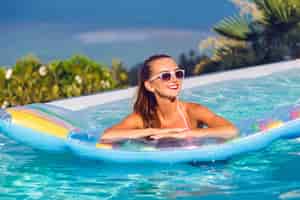 Foto gratuita retrato de estilo de vida al aire libre de impresionante joven divirtiéndose en la piscina infinita con impresionantes vistas a la isla tropical, vistiendo bikini brillante y gafas de sol, nadando en un colchón de aire.