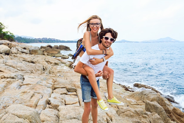 Retrato de estilo de vida al aire libre de la joven hermosa pareja de enamorados posando y divirtiéndose en la bonita playa de piedra, colores suaves.