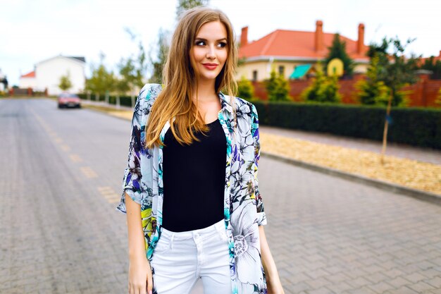 Retrato de estilo de vida al aire libre de mujer bonita rubia, pantalón blanco, camisa floral de moda, maquillaje natural pelos largos, posando en el campo, estilo callejero.