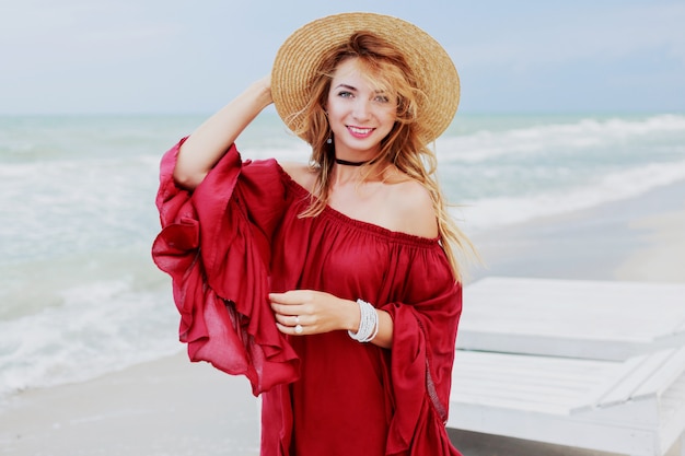 Retrato de estilo de vida al aire libre de mujer de jengibre bastante blanca en elegante vestido posando en la playa cerca del océano. Cielo azul. Clima ventoso.