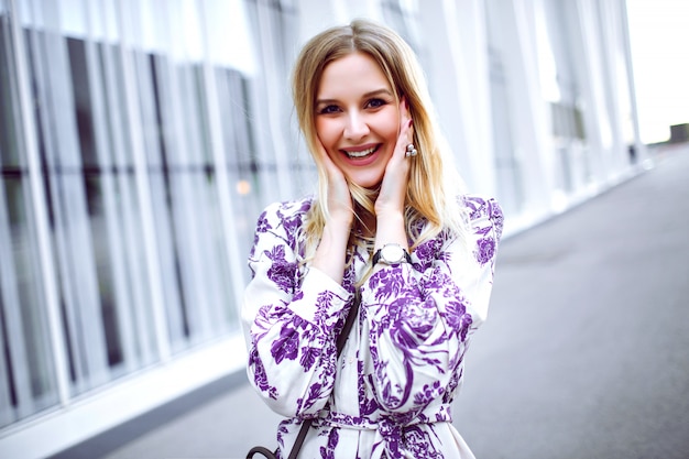 Retrato de estilo de vida al aire libre de mujer rubia sonriente emocionada con elegante vestido floral de moda y mirando a puerta cerrada, primavera verano.