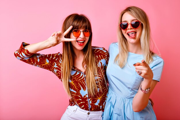 Retrato de estilo de vida de dos hermanas amigas bonitas felices, posando y divirtiéndose juntas en la pared rosa, mostrando la lengua larga y el gesto v, humor positivo de fiesta.