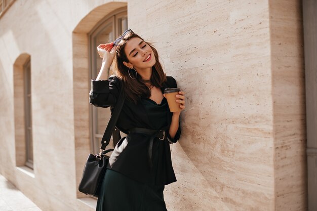 Retrato de estilo de vida de la encantadora joven morena con maquillaje brillante, traje oscuro con vestido, chaqueta, bolso, gafas de sol y cinturón de pie cerca de la pared beige al aire libre y sonriendo