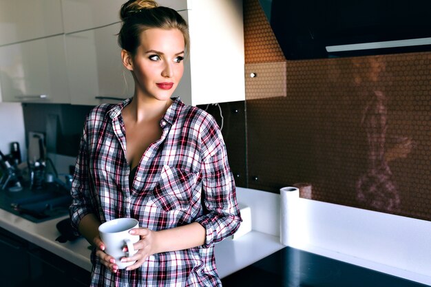 Retrato de estilo de vida interior de una mujer bonita rubia relajada y disfrutando de su tiempo de la mañana, posando en la cocina, vistiendo una acogedora camisa a cuadros, colores suaves de película vintage. bebiendo café sabroso.