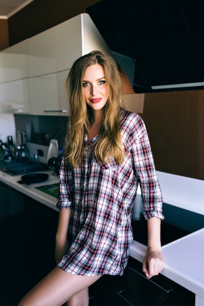 Retrato de estilo de vida interior de una mujer bonita rubia relajada y disfrutando de su tiempo de la mañana, posando en la cocina, vistiendo una acogedora camisa a cuadros, colores suaves de película vintage.