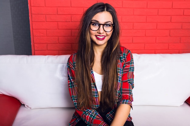 Retrato de estilo de vida de moda interior de joven bonita hipster con maquillaje brillante y sonrisa increíble, tiene pelos largos morenos, vistiendo camisa a cuadros