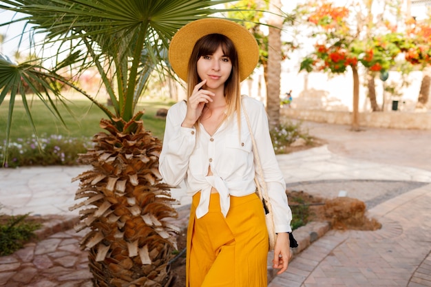 Retrato de estilo de vida de una mujer caucásica bonita con sombrero de paja, blusa blanca y bolso estilo Bali caminando en un jardín tropical.