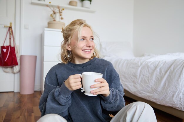 Retrato de estilo de vida de una mujer joven sentada en el suelo del dormitorio con una taza de té bebiendo de un gran blanco