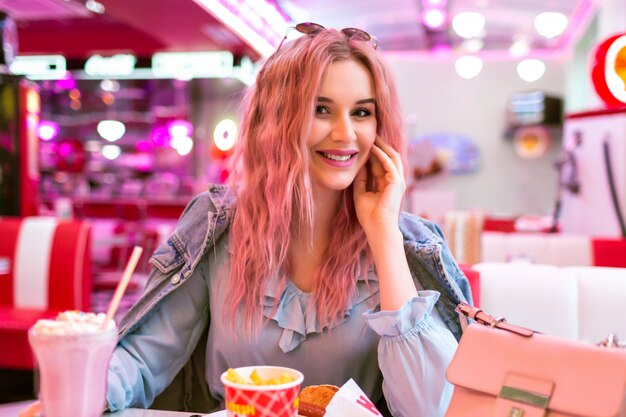 Retrato de estilo de vida positivo de una mujer bonita feliz con pelos rosados cenando en un café americano vintage, comiendo hot dog, papas fritas y mil shake, comida chatarra, colores pastel.