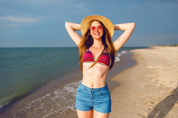 Retrato de estilo de vida positivo de una mujer joven y bonita que disfruta de sus vacaciones cerca del mar, playa solitaria alrededor, vibraciones de viaje, cuerpo delgado y saludable, sombrero de bikini y gafas de sol