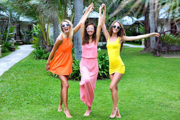 Retrato de estilo de vida tropical de verano de tres mejores amigas felices divirtiéndose al aire libre, vistiendo coloridos vestidos sexy, estilo de playa de fiesta de vacaciones, jardín exótico, ropa de moda, gafas de sol, relax, alegría