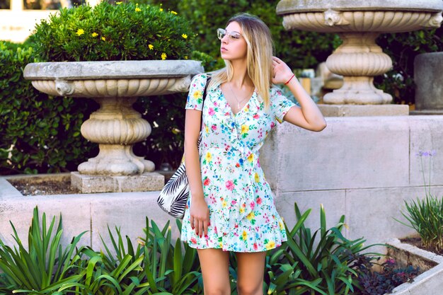 Retrato de estilo de vida de verano de feliz magnífica mujer rubia elegante con vestido de té floral de moda elegante ligero y posando en el viejo parque europeo, humor de viaje.