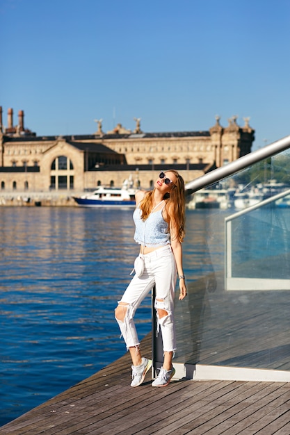 Retrato de estilo de vida de verano de mujer bonita rubia viaja sola en Barcelona, hermosa arquitectura y vista al mar, look de estilo callejero de moda, vacaciones, alegría, viajero, top corto y denim.