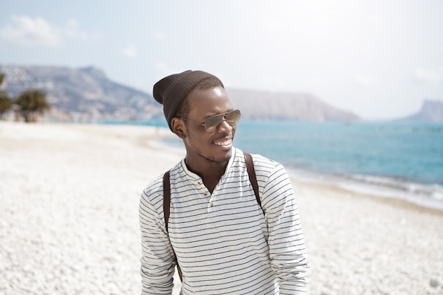 Retrato de estilo de vida de verano del viajero de moda joven de piel oscura con mochila caminando en la playa soleada durante las vacaciones, admirando el pintoresco paisaje y el mar en calma con una sonrisa alegre y feliz