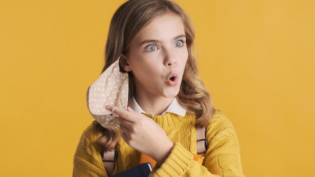 Retrato de una estudiante adolescente rubia quitándose la máscara protectora sintiendo alivio después del día de estudio sobre fondo amarillo El primer concepto de seguridad