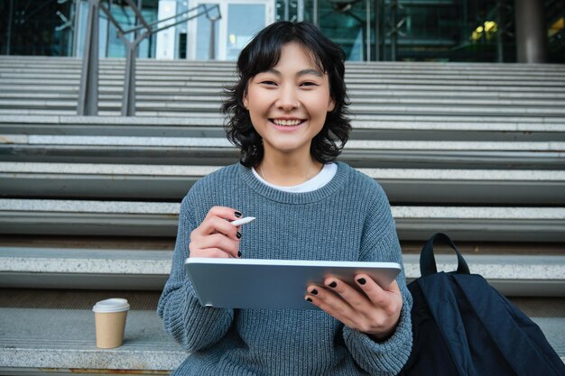 Retrato de una estudiante asiática hipster sentada en las escaleras con una tableta digital y una taza de café dibuja