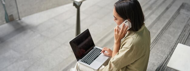 Foto gratuita retrato de una estudiante asiática sentada en las escaleras con una computadora portátil hablando por teléfono móvil una joven hace un