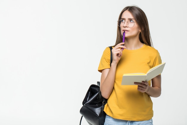 Retrato de una estudiante casual sonriente con mochila escribiendo en un bloc de notas mientras está de pie con libros aislados sobre la pared blanca