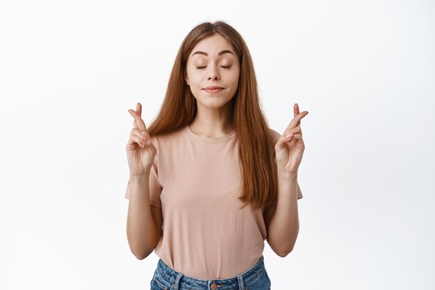 Retrato de una estudiante esperanzada que desea que el examen cruce los dedos para tener buena suerte y cerrar los ojos rezando suplicando que el sueño se haga realidad fondo blanco