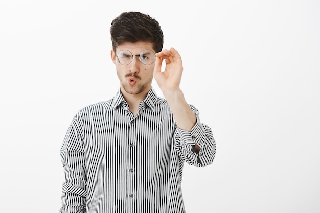 Retrato de un estudiante europeo adulto ordinario, quitándose las gafas y limpiando las gafas, mirando las gafas enfocadas, doblando los labios para soplar, de pie sobre una pared gris