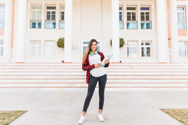 Retrato de un estudiante femenino que sostiene los libros en la mano que se coloca delante de universidad