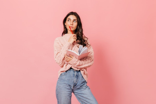 Retrato de estudiante inteligente con cuaderno y lápiz rojo mirando pensativamente a la cámara sobre fondo rosa.