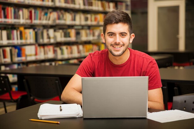 Retrato de un estudiante latino atractivo que hace un trabajo escolar con una computadora portátil en la biblioteca