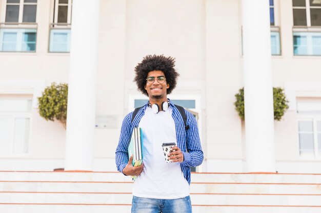 Retrato de un estudiante masculino adolescente afro feliz que sostiene los libros y la taza de café para llevar que se coloca delante de universidad