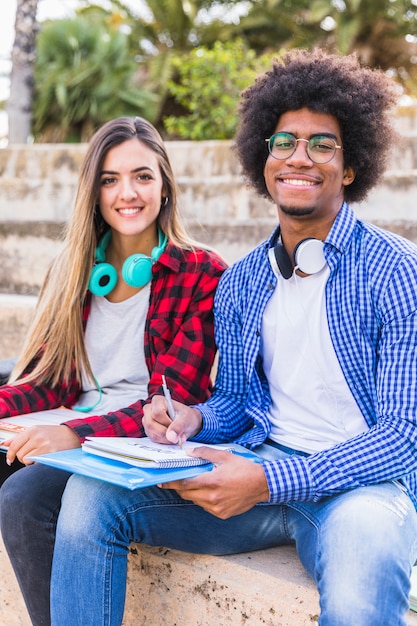 Retrato del estudiante masculino afro sonriente que se sienta con su novia en el aire libre con los libros