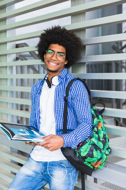 Foto gratuita retrato de un estudiante masculino afro sonriente sosteniendo el libro en la mano apoyado contra la pared
