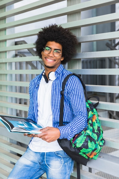 Retrato de un estudiante masculino afro sonriente sosteniendo el libro en la mano apoyado contra la pared