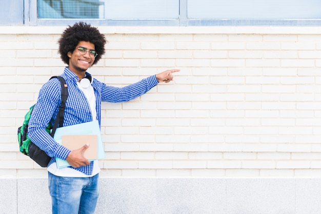 Retrato de un estudiante masculino sonriente africano que sostiene los libros en la mano que señala el dedo en la pared pintada blanca