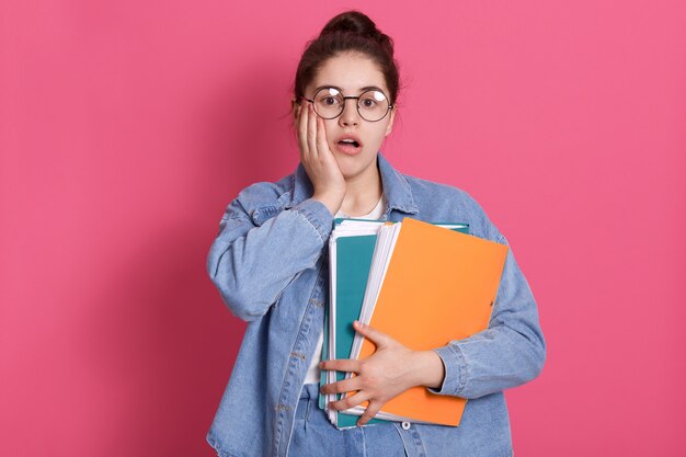 Retrato de estudiante con moño, usa jeans y gafas redondas, sostiene carpetas de papel de colores
