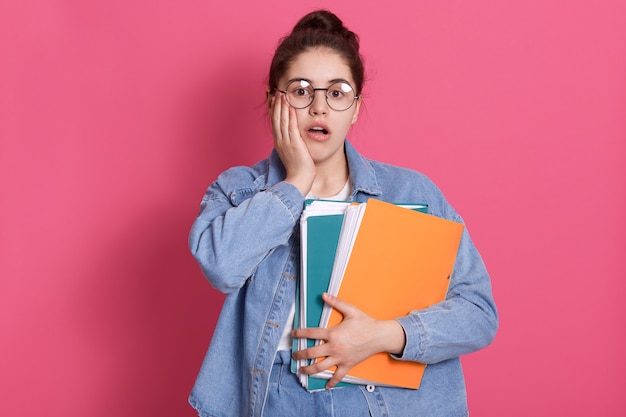 Retrato de estudiante con moño, usa jeans y gafas redondas, sostiene carpetas de papel de colores