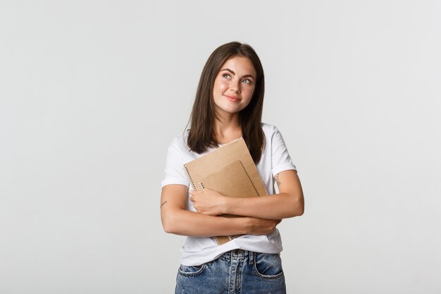 Retrato de una estudiante nostálgica sonriente mirando la esquina superior izquierda, sosteniendo cuadernos.