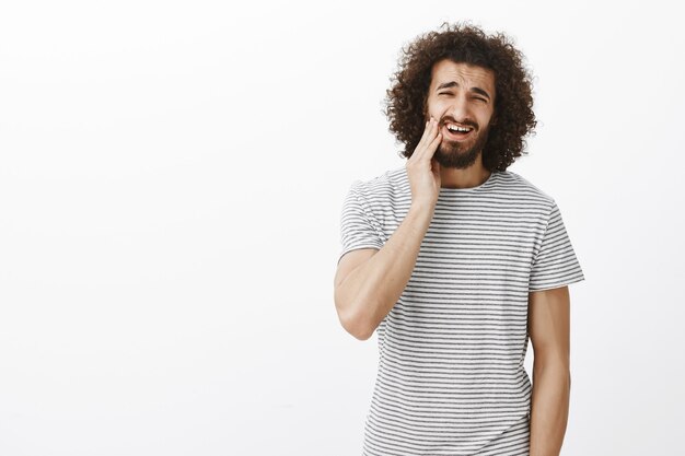 Retrato de un estudiante oriental guapo molesto disgustado con peinado afro en camiseta a rayas, tocando la barba y haciendo muecas de disgusto, sintiendo la necesidad de afeitarse la barba