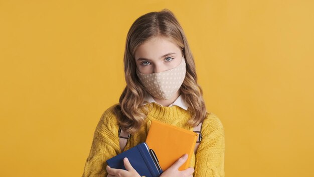 Retrato de una estudiante rubia adolescente con una máscara protectora parada con libros de texto que se ven serios aislados en un fondo amarillo. Seguridad primero