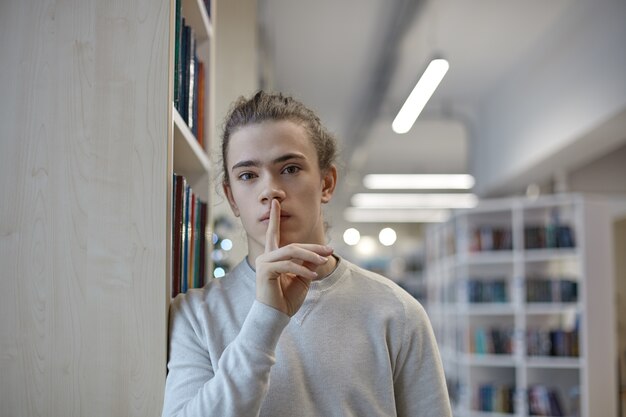 Retrato de estudiante de secundaria serio en ropa casual de pie en la biblioteca y mostrando gesto de silencio