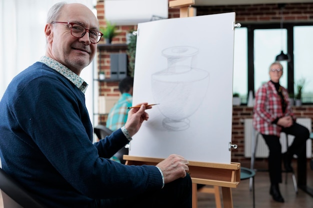 Retrato de un estudiante senior sonriente dibujando un modelo de jarrón usando un lápiz gráfico trabajando en un boceto durante la lección de arte desarrollando habilidades artísticas para el crecimiento personal. Resoluciones de Año Nuevo