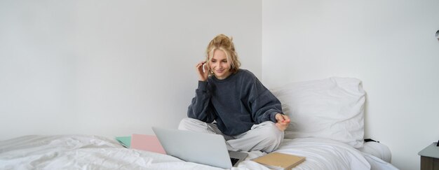 Retrato de una estudiante sentada en la cama con una computadora portátil y cuadernos estudiando remotamente en línea