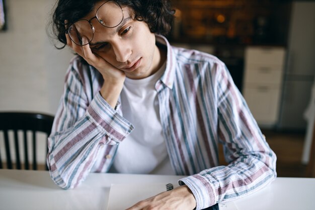 Retrato de estudiante con sueño en ropa casual sentado en el escritorio blanco con la mano en la cara, con mirada aburrida, cansado de hacer la tarea, necesita dormir.