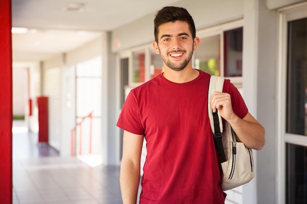 Retrato de un estudiante universitario hispano que lleva una mochila y está parado en un pasillo de la escuela