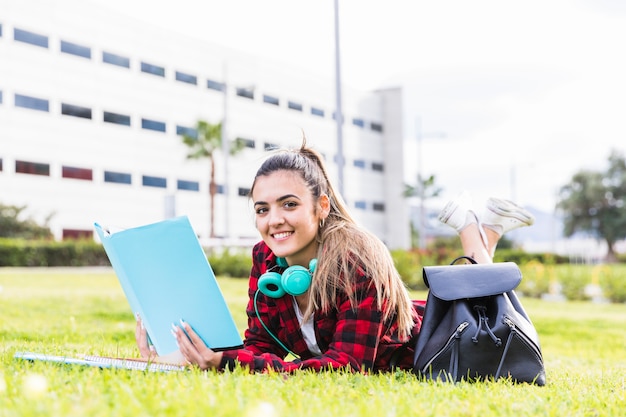 Retrato del estudiante universitario de sexo femenino sonriente que miente en la hierba verde que sostiene el libro disponible