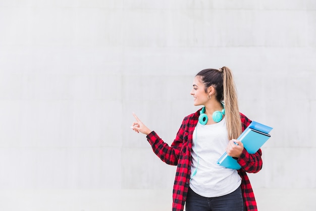 Retrato de un estudiante universitario de sexo femenino sonriente que sostiene los libros en la mano que señala su dedo en la pared blanca con el espacio de la copia
