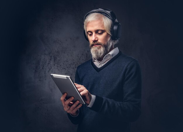 Retrato de estudio de un apuesto anciano que usa una tableta con auriculares sobre un fondo oscuro.