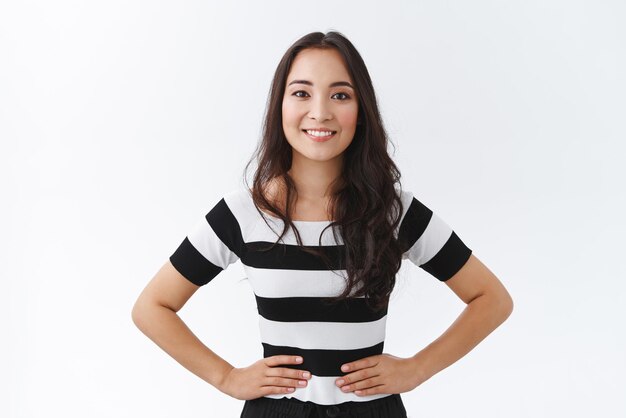 Retrato de estudio atractiva y animada mujer morena asiática con camiseta a rayas cogida de la mano en las caderas de pie confiada y optimista sonriendo ampliamente mostrando una determinada actitud segura de sí misma