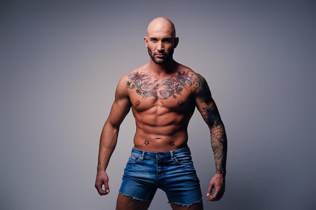 Retrato de estudio de cabeza afeitada sin camisa, hombre musculoso con tatuajes en el torso vestido con pantalones cortos de mezclilla sobre fondo de viñeta gris.