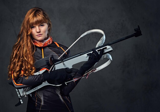 Retrato de estudio de una campeona de biatlón pelirroja sostiene un arma sobre fondo gris.