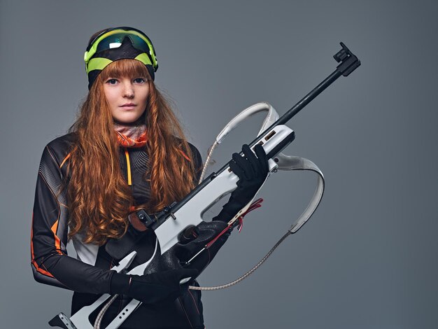 Foto gratuita retrato de estudio de una campeona de biatlón pelirroja sostiene un arma sobre fondo gris.