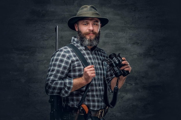 Retrato de estudio de un cazador barbudo que lleva una camisa de lana a cuadros sostiene un rifle.